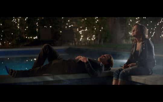 Harry laying besides a pool talking to Daisy who has her feet in the pool.