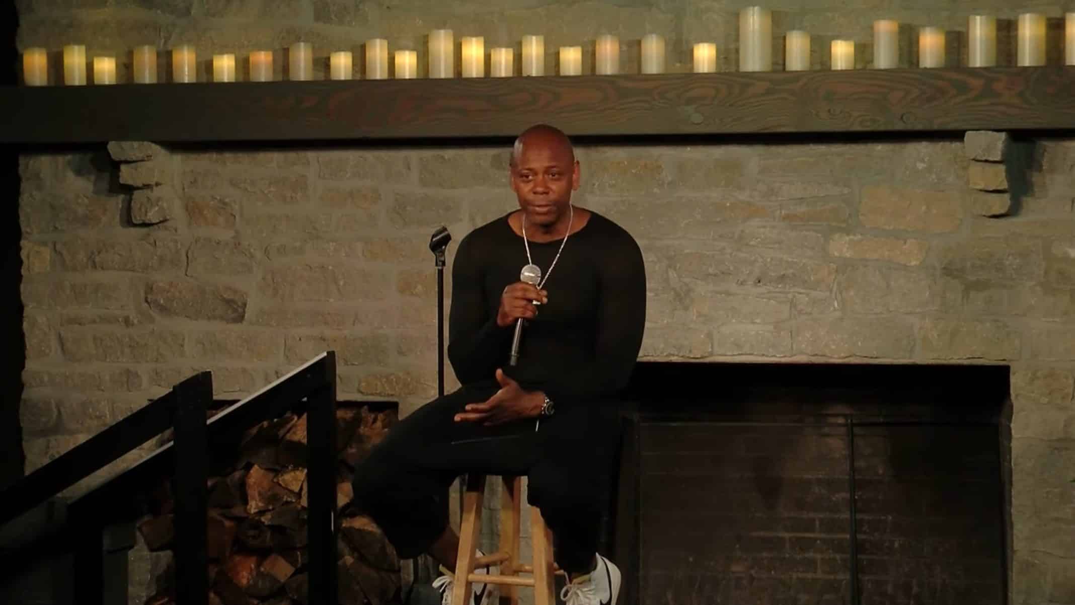 Dave Chappelle sitting on a stool, talking to an audience.