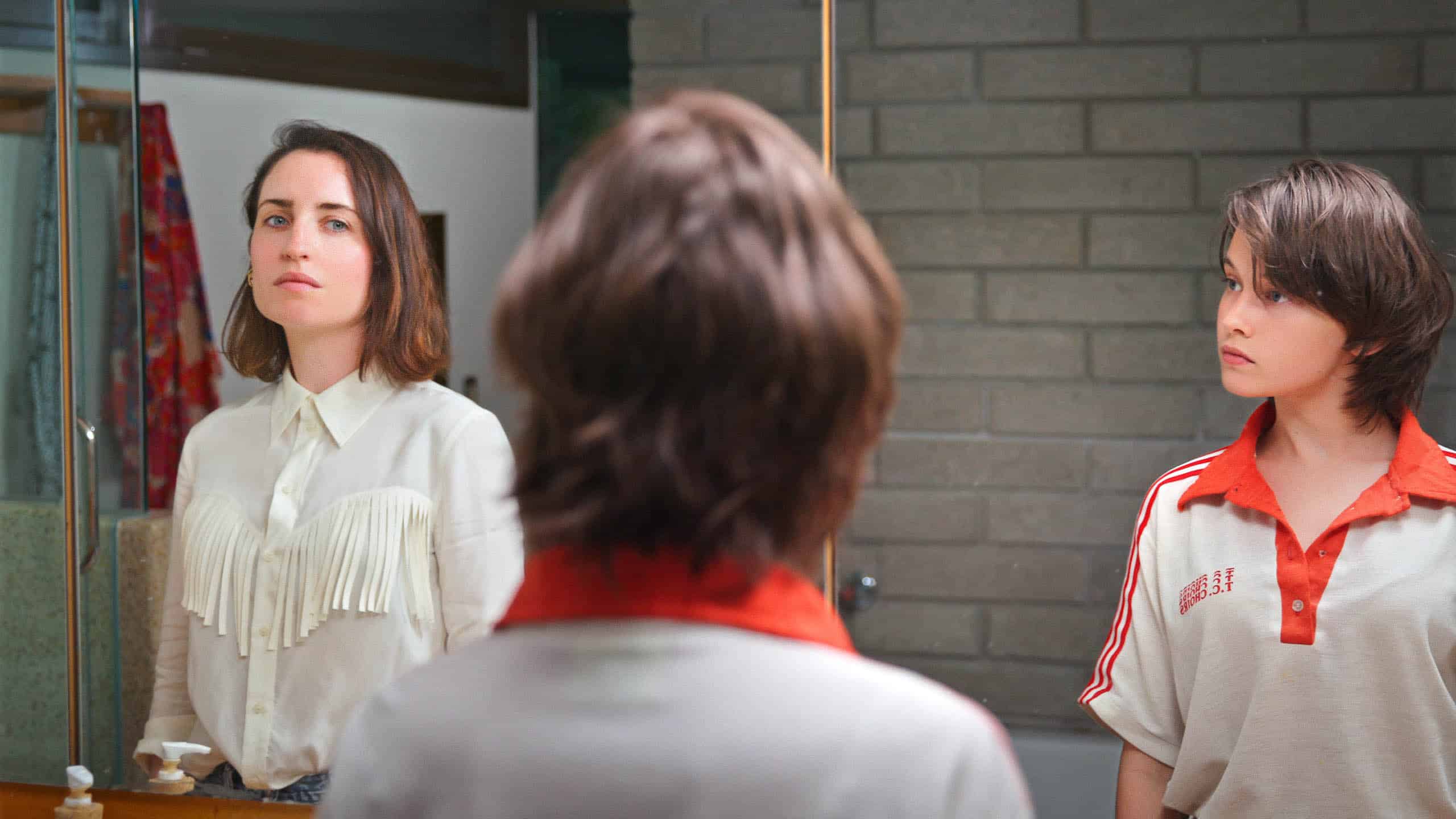 Liza (Zoe Lister-Jones) and Liza's Younger Self (Cailee Spaeny) both in front of Liza's bathroom mirror