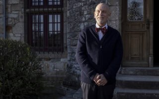 Andrew Blake (John Malkovich) standing in front of the Beauvillier estate – Mr. Blake At Your Service