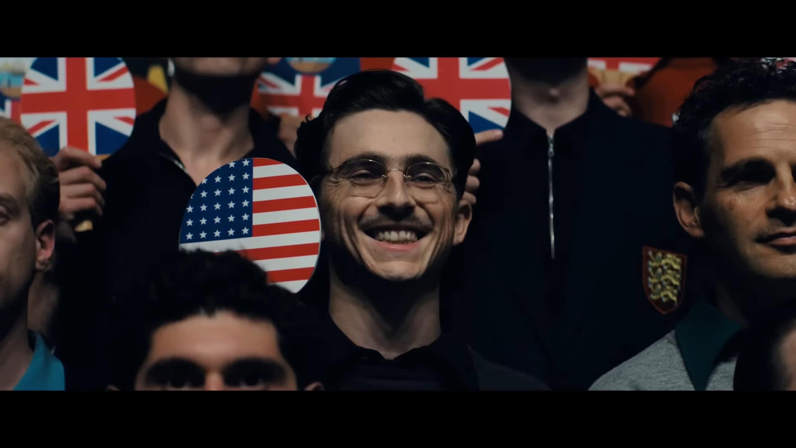 Marty (Timothée Chalamet) smiling a paddle that has the US flag on it, as he takes a group photo at the British Open in Marty Supreme 